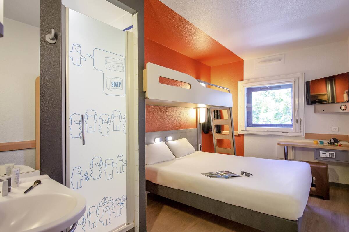 Une chambre d’hôtel avec un lit superposé, une table de chevet, une fenêtre, un lavabo et un miroir.
