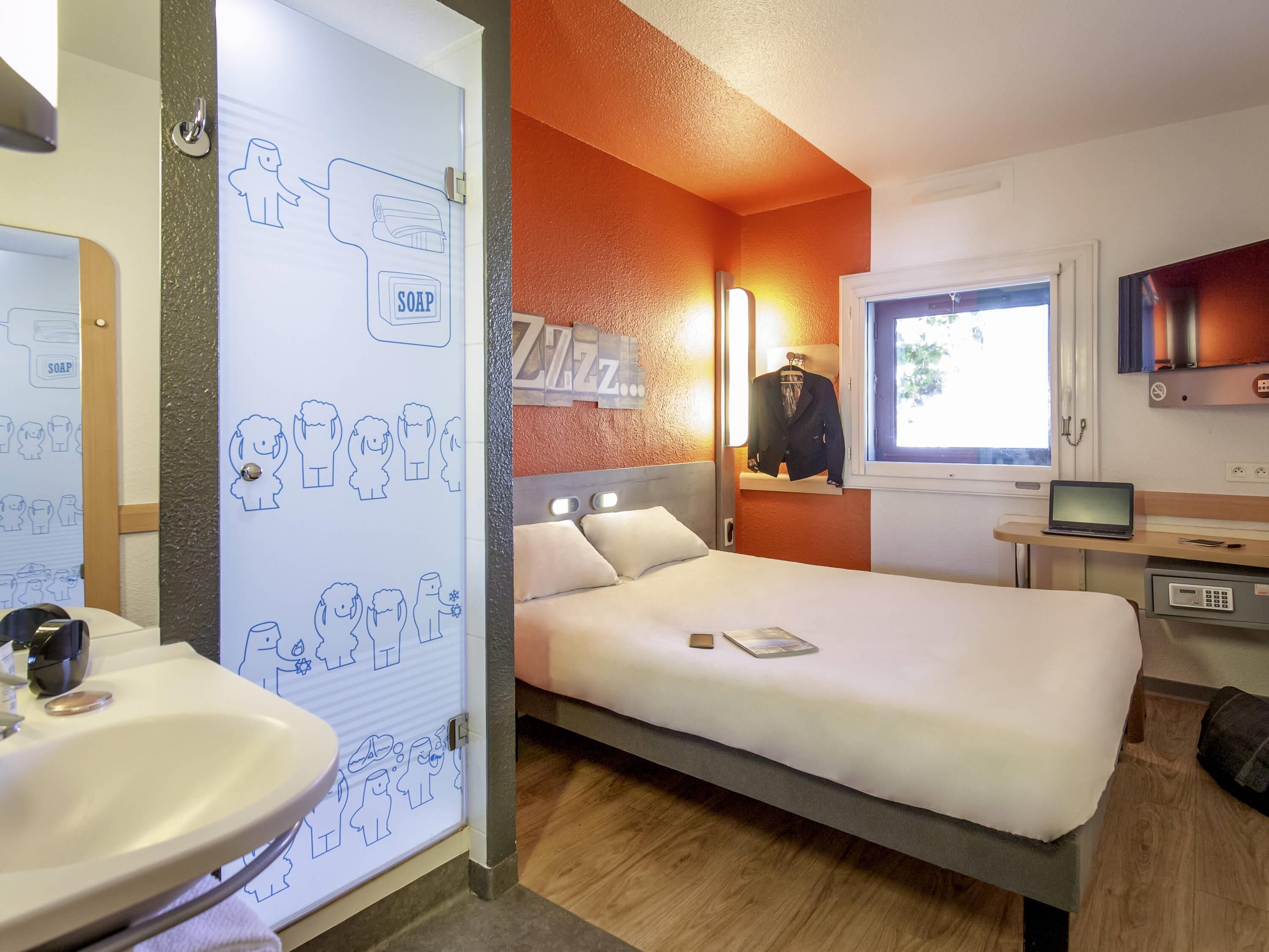 A hotel room with a bed, a bedside table with a laptop, a mirror, a window, and a wall with a soap dispenser.