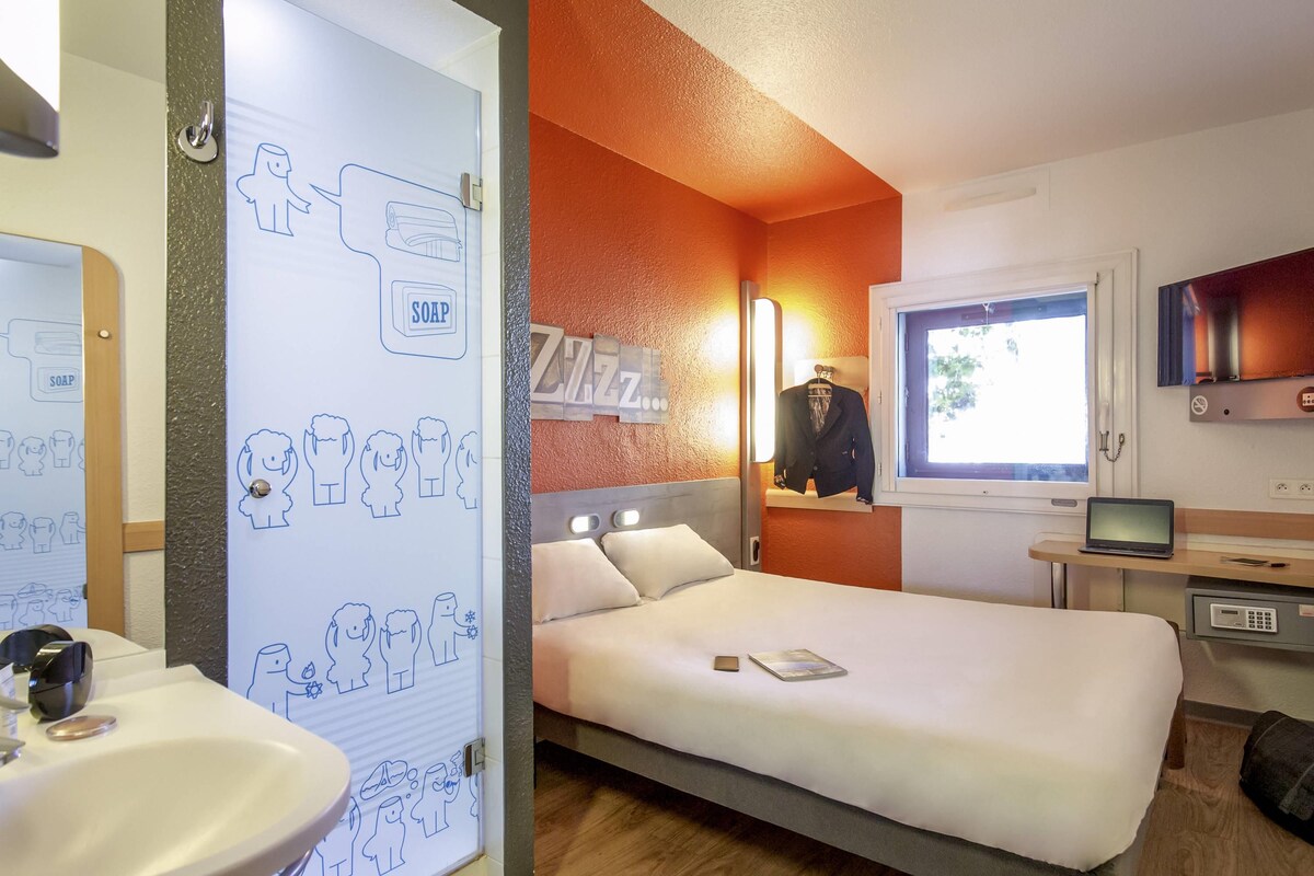 Une chambre d’hôtel avec un lit, une table de chevet sur laquelle se trouve un ordinateur portable, un miroir, une fenêtre et un mur muni d’un distributeur de savon.