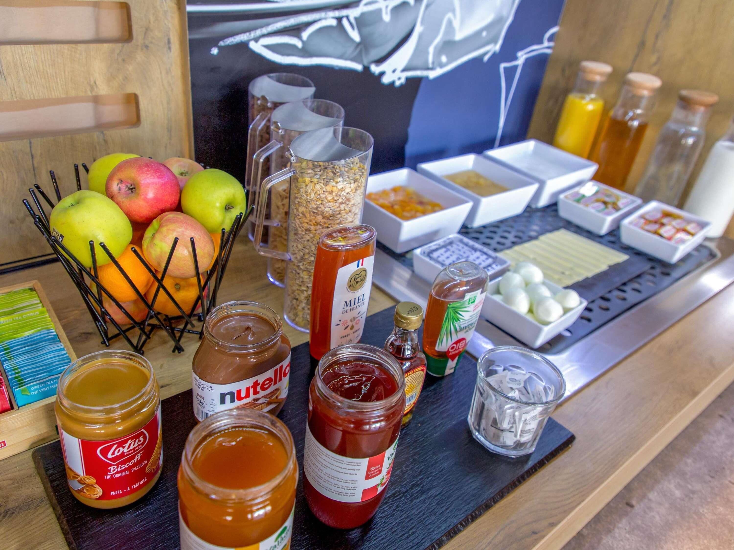 Una mesa con diferentes alimentos, incluyendo frascos de Nutella, miel y mermelada, además de frutas y condimentos.