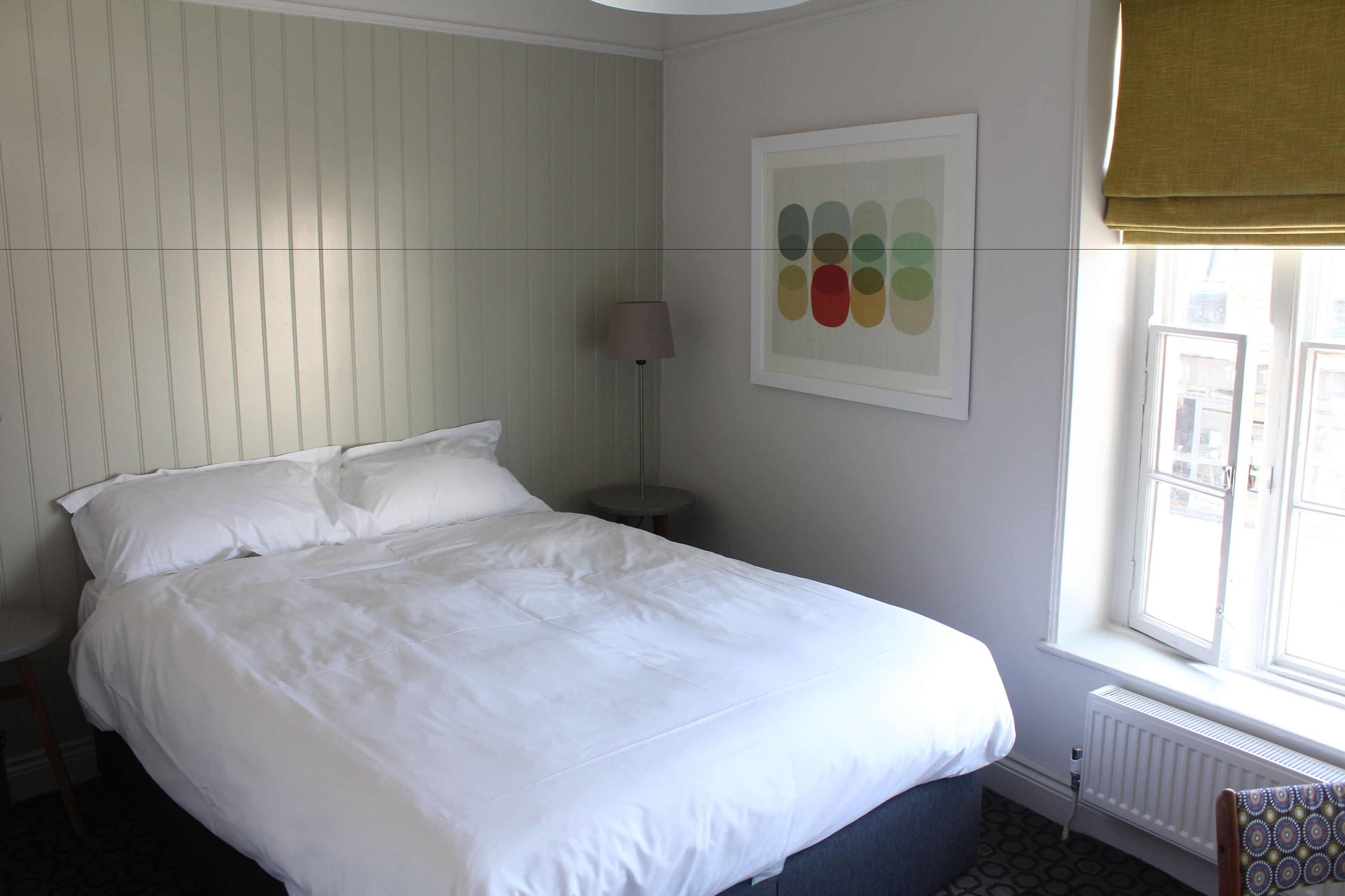 A neatly made bed in a room with a radiator, a lamp, and a window with a view of buildings.