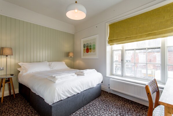 A neatly made bed with white linens, a round table with a lamp, and a window with a view of buildings. - The Dolphin (Newbury)