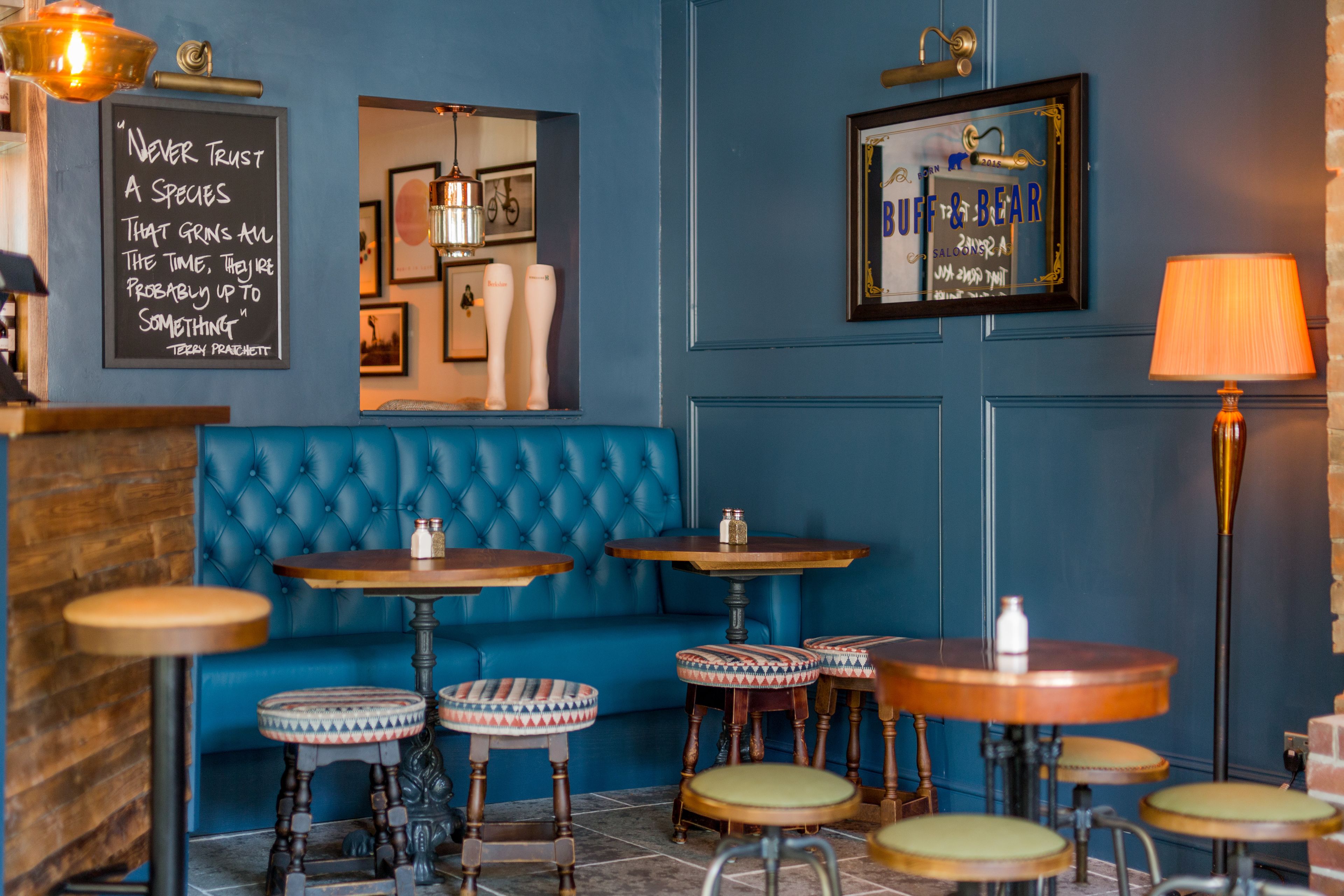 A cozy bar area with blue walls, a tufted blue sofa, and several round wooden tables with stools.