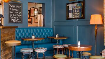 Un rincón de bar acogedor con paredes azules, un sofá azul con capitoné y varias mesas redondas de madera con taburetes.