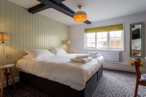 A neatly made bed with white linens and towels, a bedside table with a lamp, a window with a yellow blind, and a mirror on the wall.