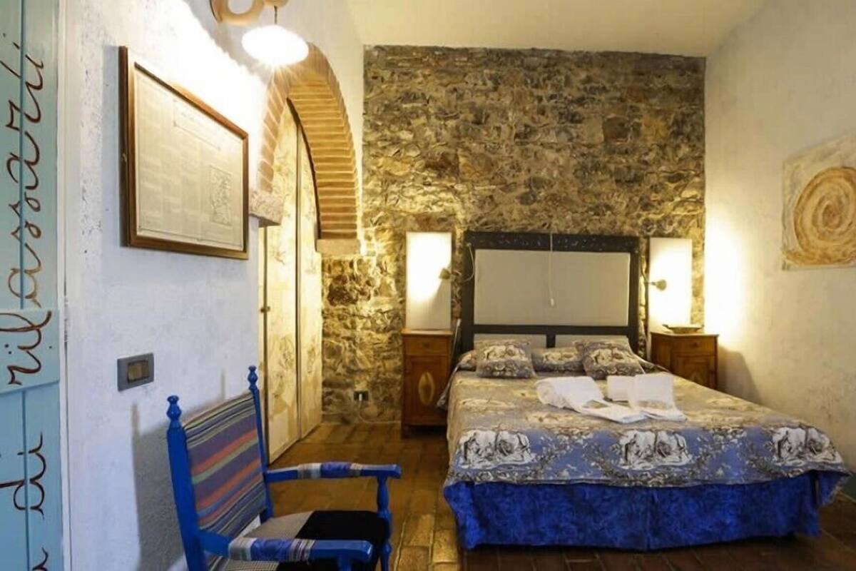 A bedroom with a stone wall, a bed with a blue bedspread, a wooden nightstand, a blue chair, and a framed picture on the wall.