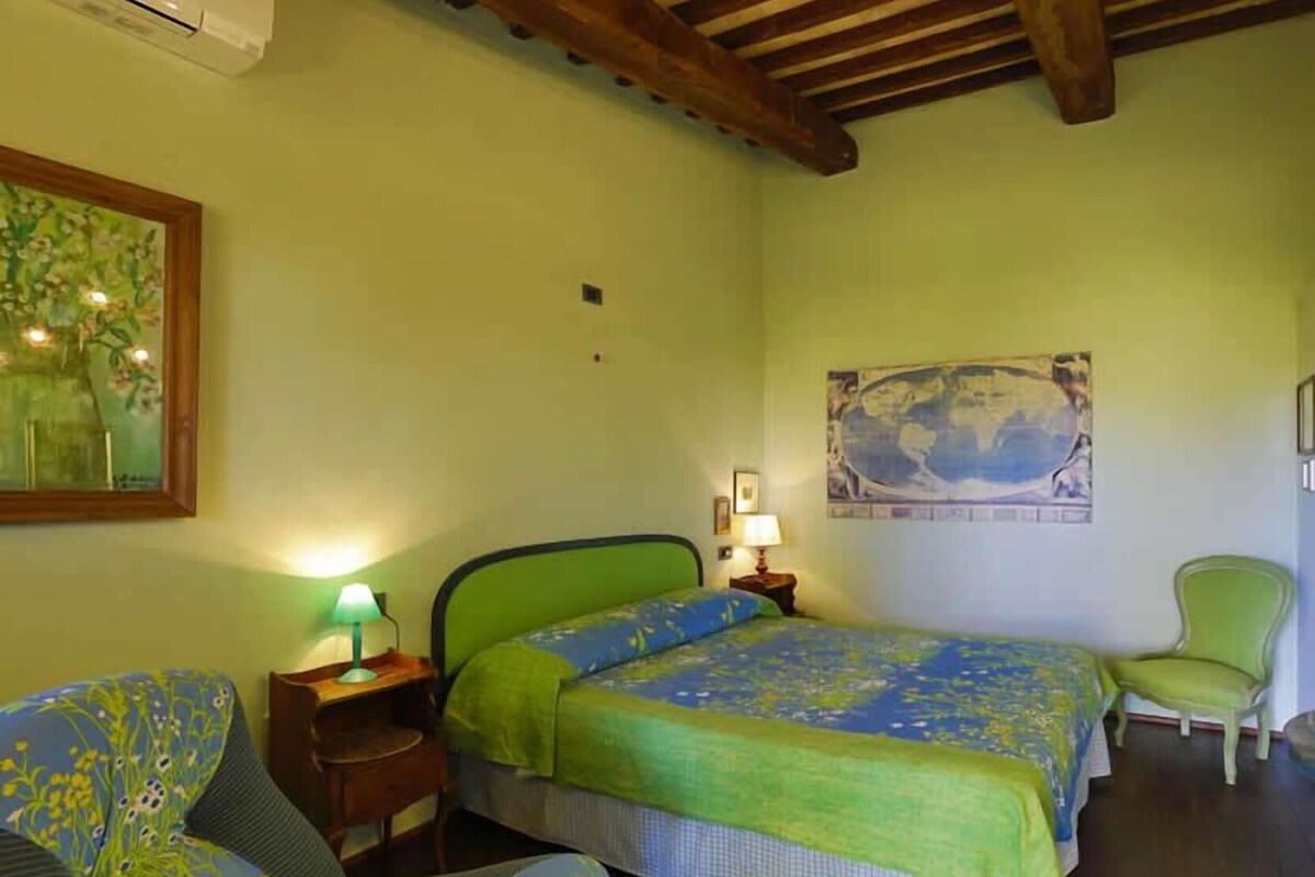 A bedroom with a bed, a chair, a nightstand, a lamp, a painting, and an air conditioning unit.