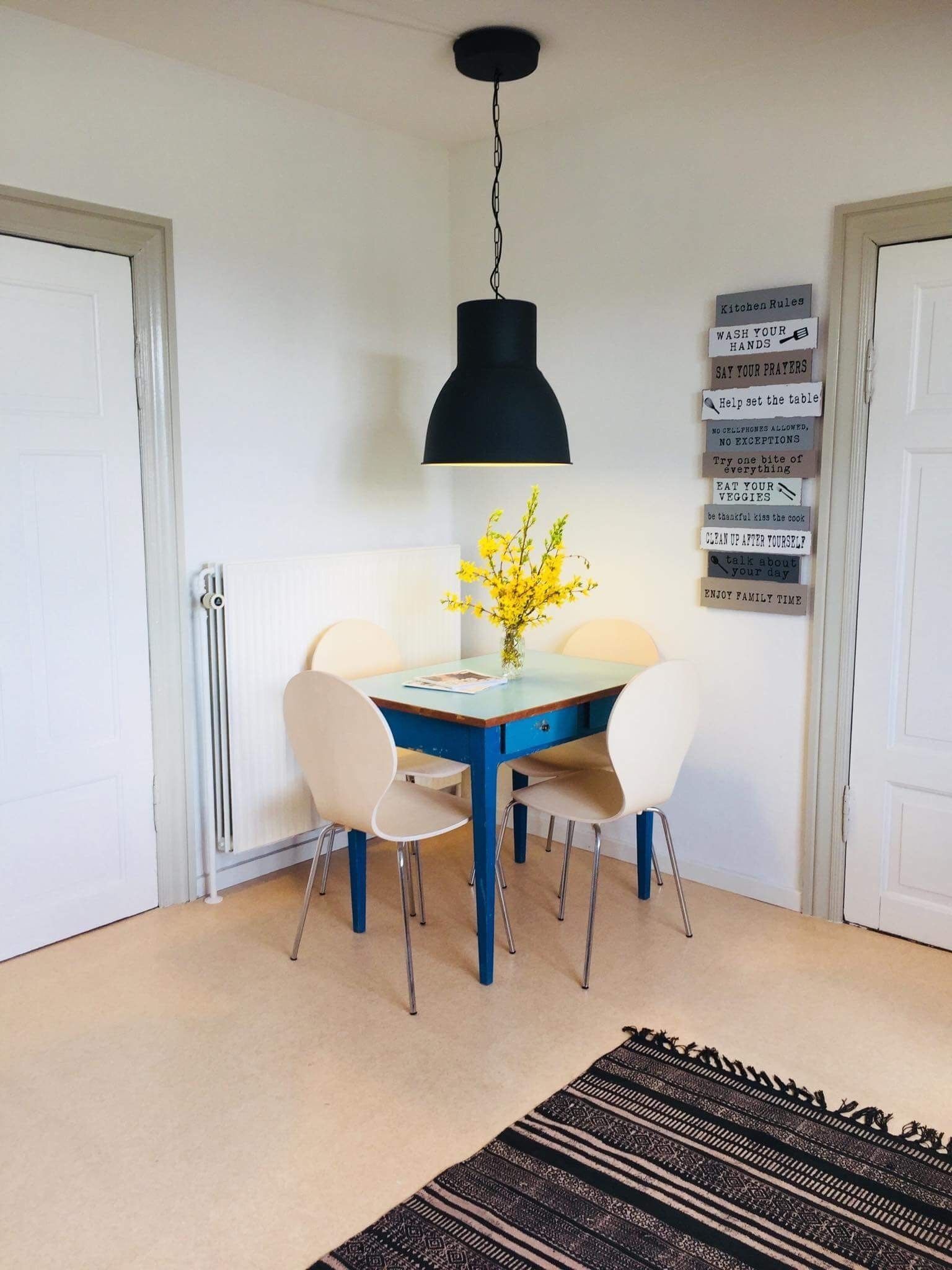 Uma sala de jantar com uma mesa azul, cadeiras brancas, uma luminária pendente preta e um letreiro na parede com regras da cozinha.