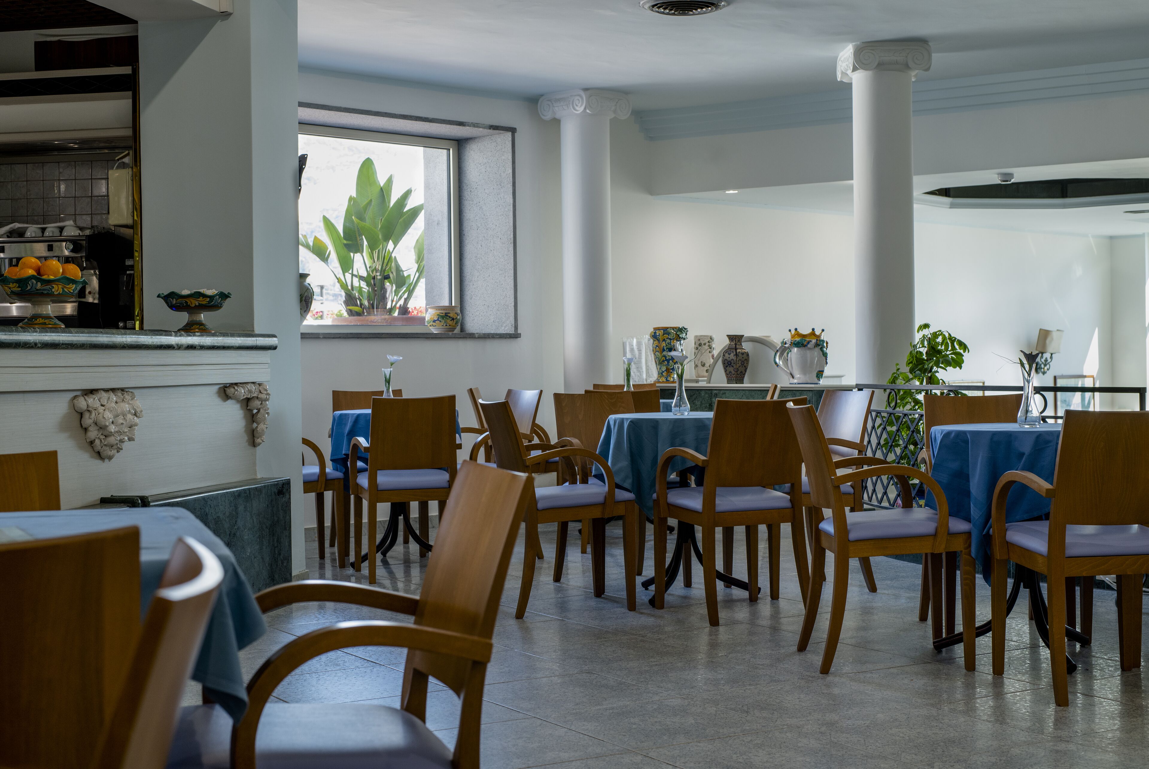 A dining area with wooden chairs and tables, a kitchen in the background, and a window with plants.