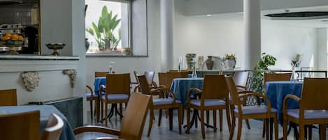 A dining area with wooden chairs and tables, a kitchen in the background, and a window with plants.