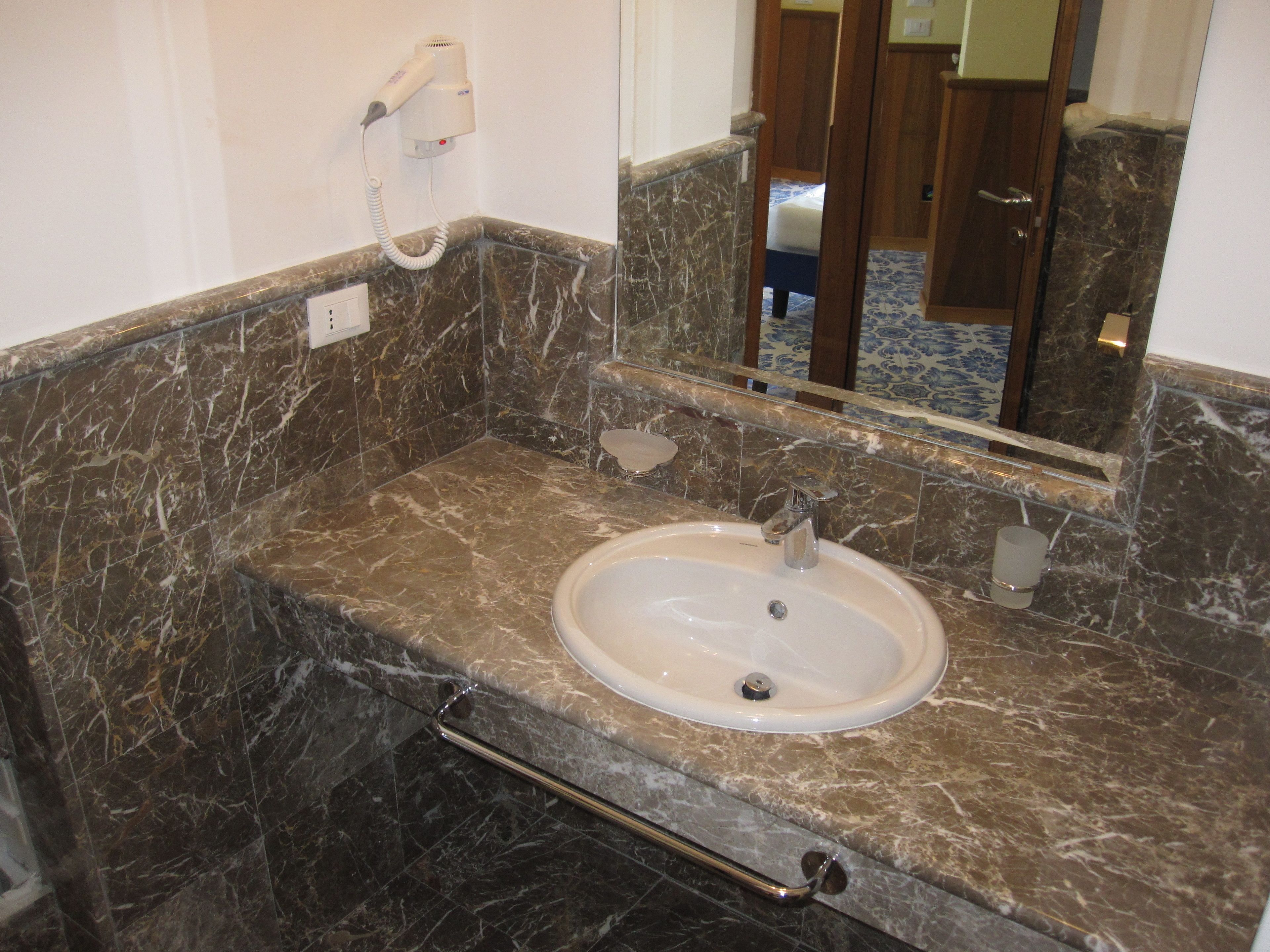 A bathroom with a marble sink, a handheld hair dryer mounted on the wall, and a mirror reflecting a room with a bed and a door.