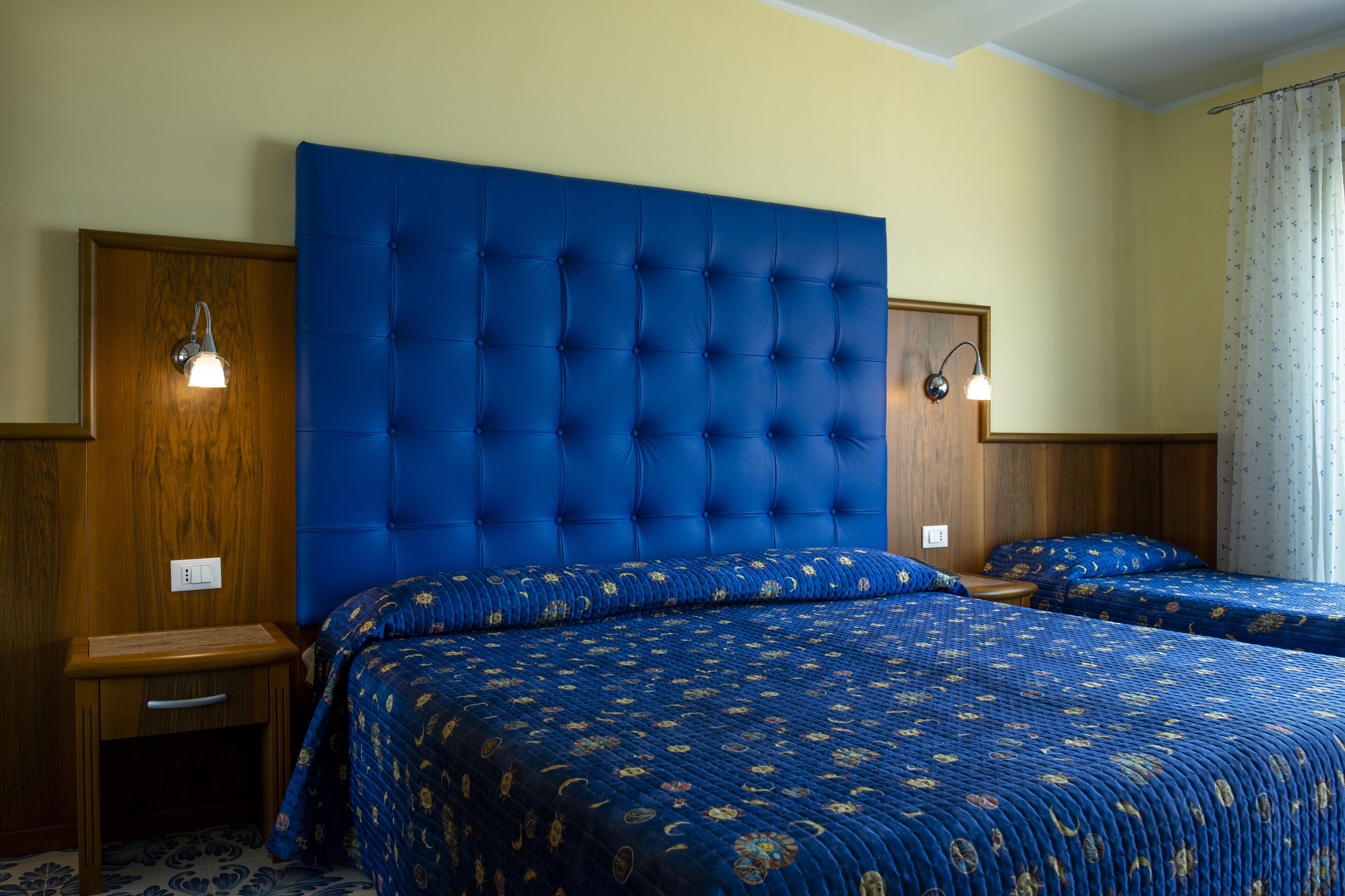 A hotel room with a large tufted headboard, two beds with matching blue floral bedding, wooden nightstands, and wall-mounted light fixtures.