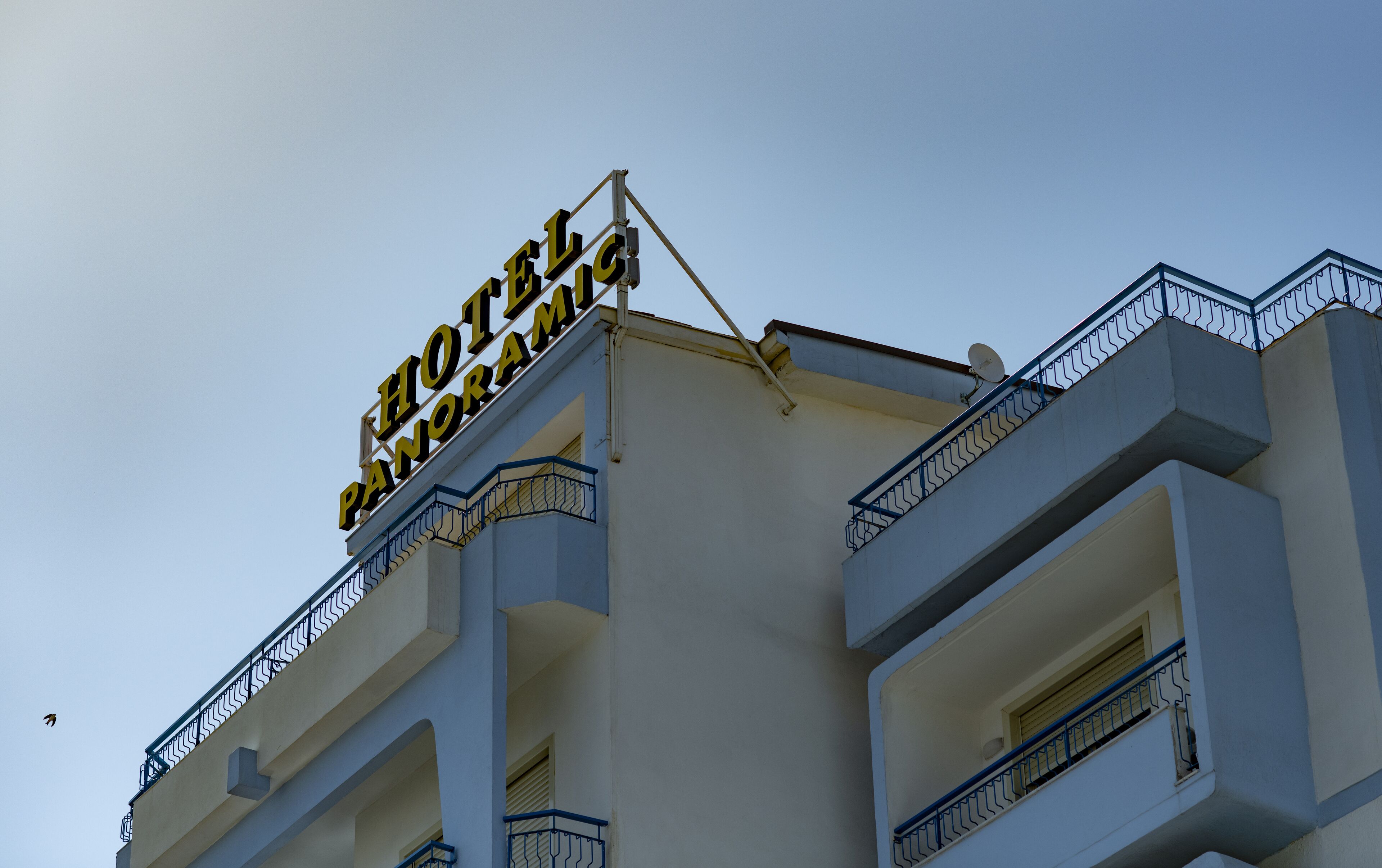 Une enseigne d’hôtel portant l’inscription « HÔTEL PANORAMA », fixée à un immeuble doté de nombreux balcons, avec un ciel dégagé en arrière-plan.