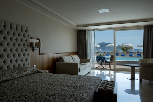 A hotel room with a large bed, a sofa, a small table, and a view of a pool and umbrellas.