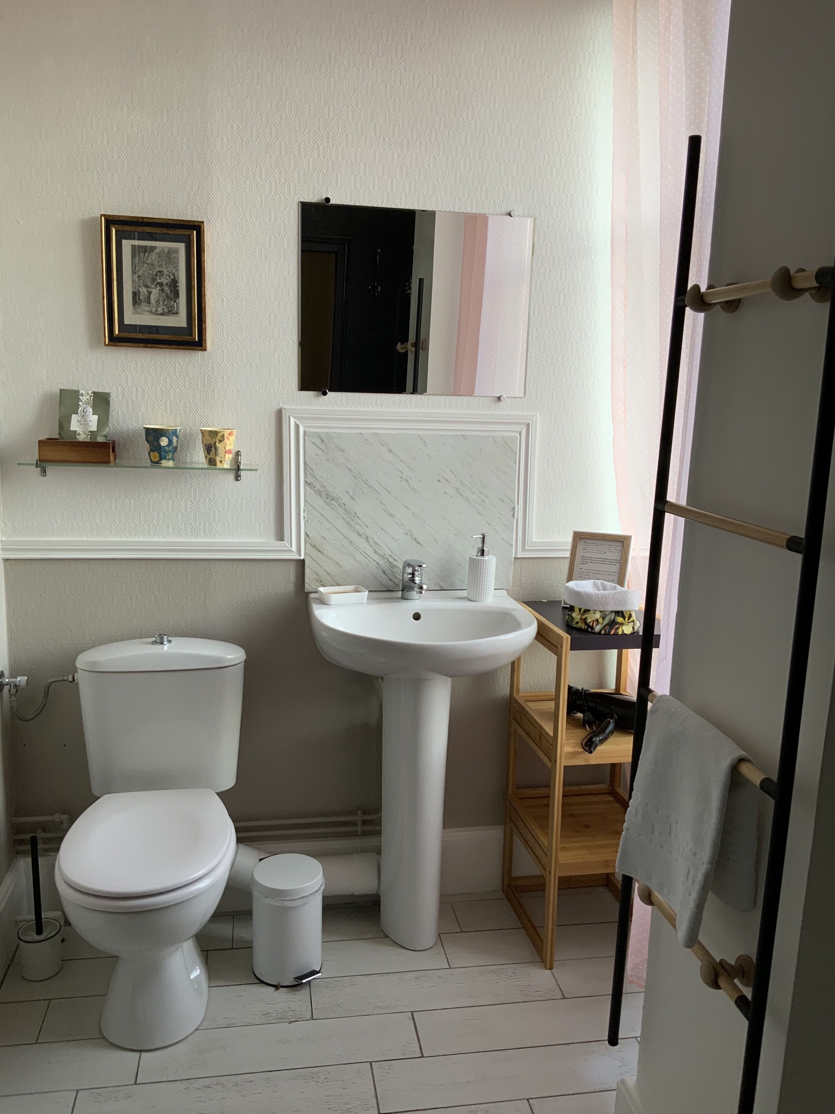 Casa de banho com sanita, lavatório e espelho sobre o lavatório. Há uma estante com objetos decorativos e um porta-retrato na parede.