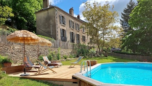 A wooden deck with lounge chairs and a thatched umbrella, a swimming pool, and a stone building in the background. - Maison Volière (La Souterraine)