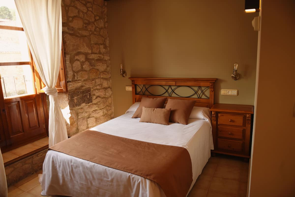 Ein Schlafzimmer mit Steinwand, Holz-Kopfteil, einem Bett mit weißer und brauner Bettwäsche, einem Nachttisch und einem Fenster mit Vorhängen.