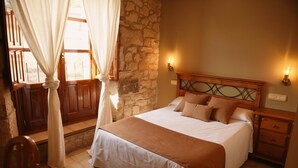Une chambre avec un mur en pierre, une tête de lit en bois et un lit recouvert d’une couverture marron. On y trouve deux lampes de chevet et une table de chevet en bois.