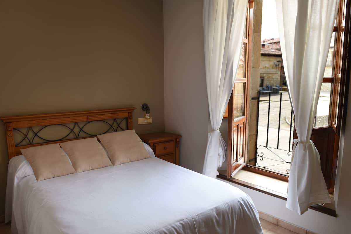 Ein Schlafzimmer mit einem hölzernen Kopfstück, einem Bett mit weißen Leinen, einem Nachttisch und einem Fenster mit Vorhängen.