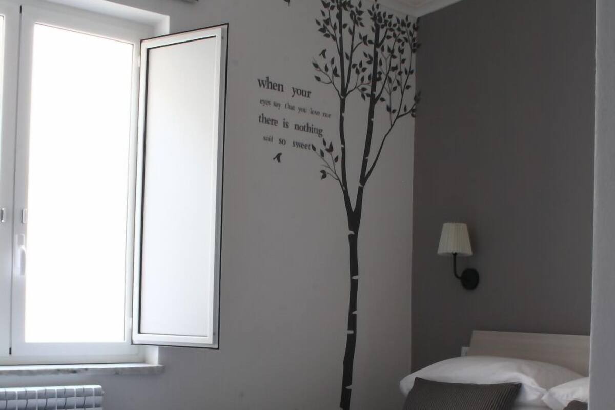 A bedroom with a bed, a window, a radiator, a wall-mounted lamp, and a tree decal.