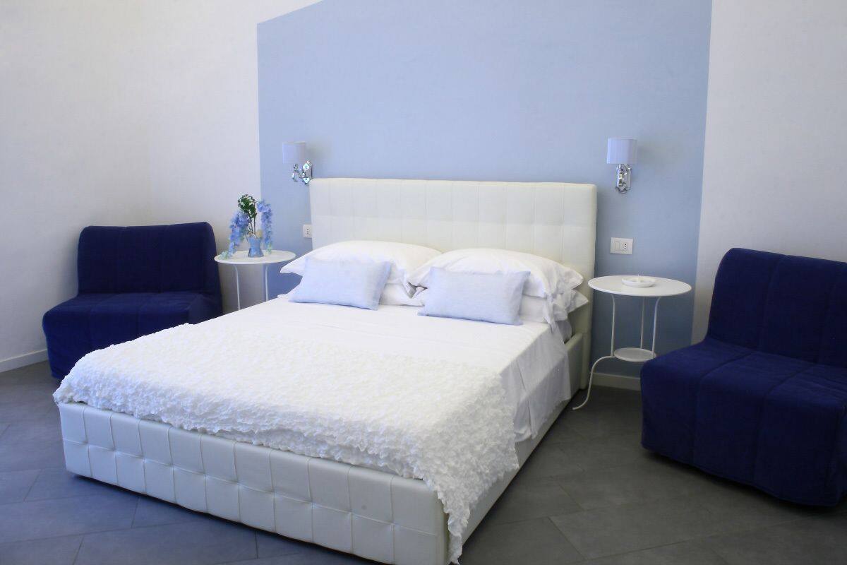 A bedroom with a white bed, two blue armchairs, a small round table, and a wall-mounted lamp.