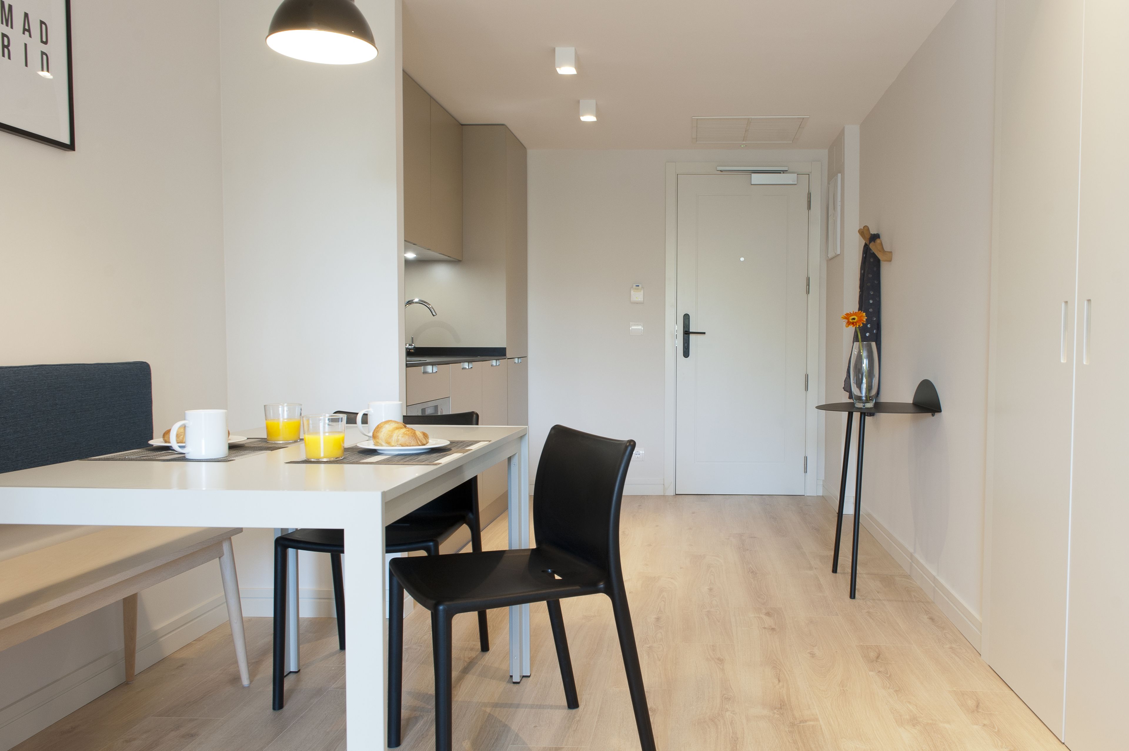 Une cuisine moderne avec un coin repas, une table blanche, des chaises noires et une petite étagère sur laquelle est posé un vase.