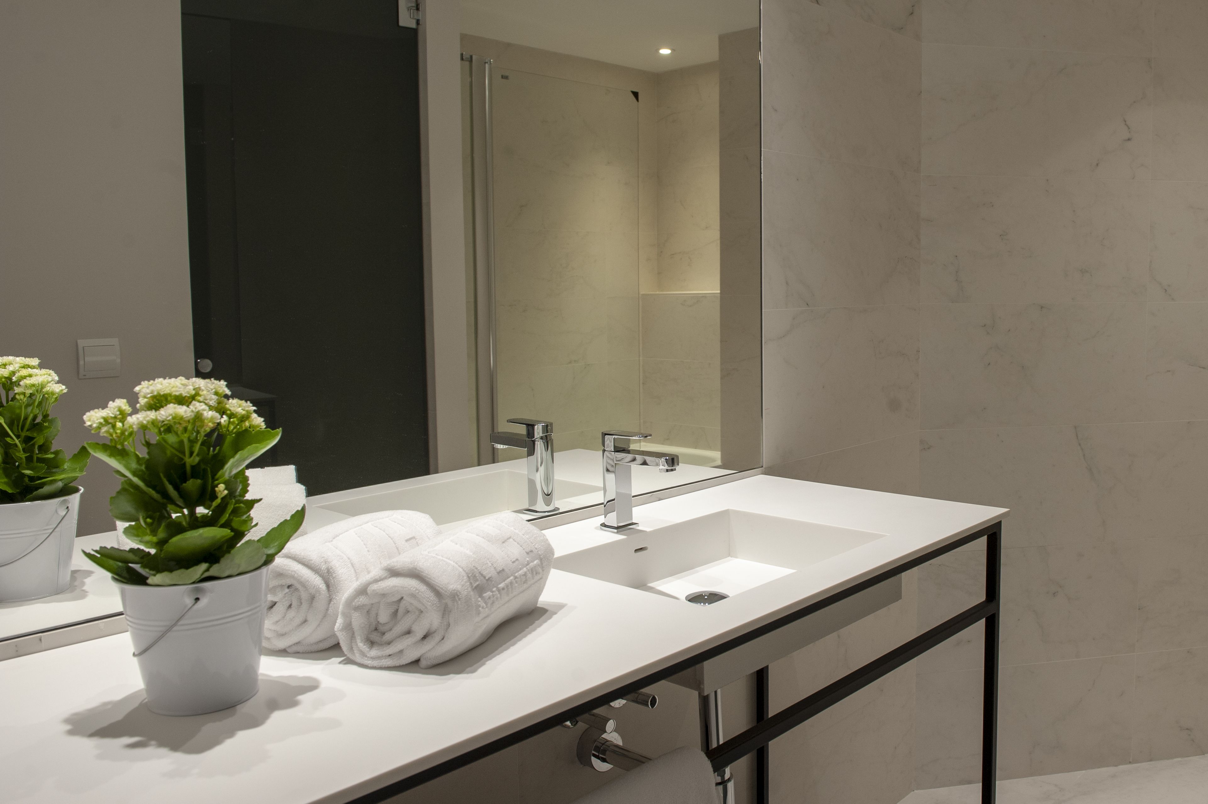 A modern bathroom with a large mirror, two sinks, and a shower. There are potted plants and neatly folded towels on the counter.