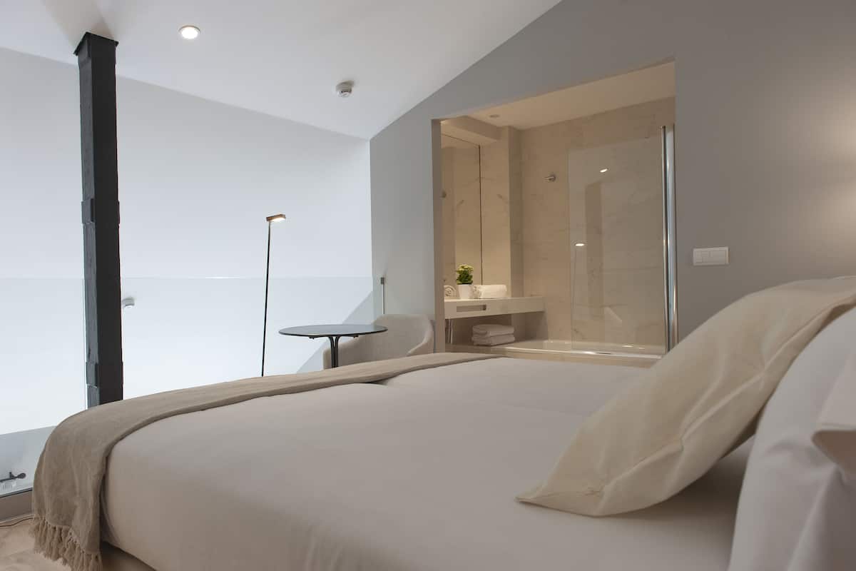 A modern hotel room with a large bed, a small table, a bathroom with a glass shower, and a view of the city.