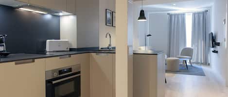 A modern kitchen with built-in appliances, a small dining area with a chair and ottoman, and a view of the living room through a doorway.