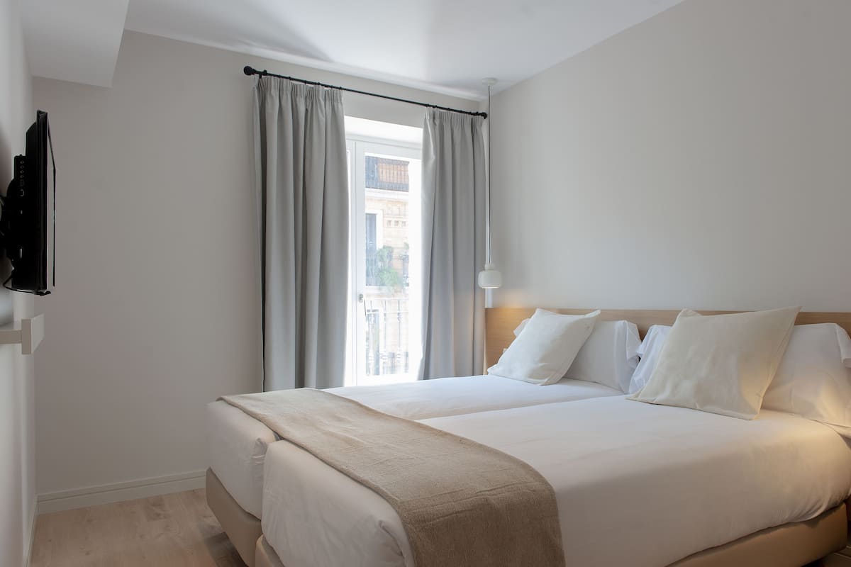 A double bed with white linens, a wooden headboard, and a view of a building through the window.