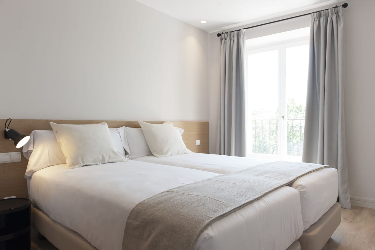 A double bed with white bedding and a grey throw blanket, a bedside lamp, and a window with grey curtains.