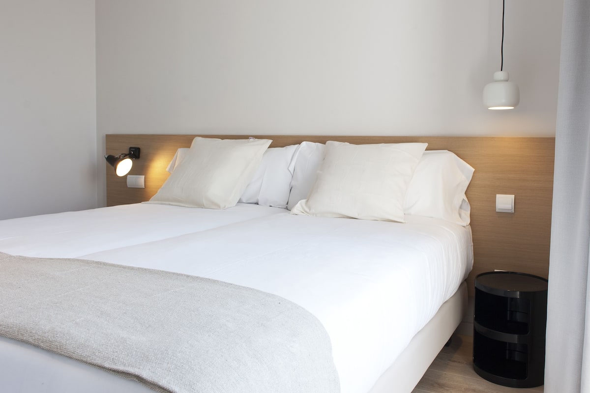 A neatly made bed with white linens and pillows, a bedside lamp, and a small black trash bin.