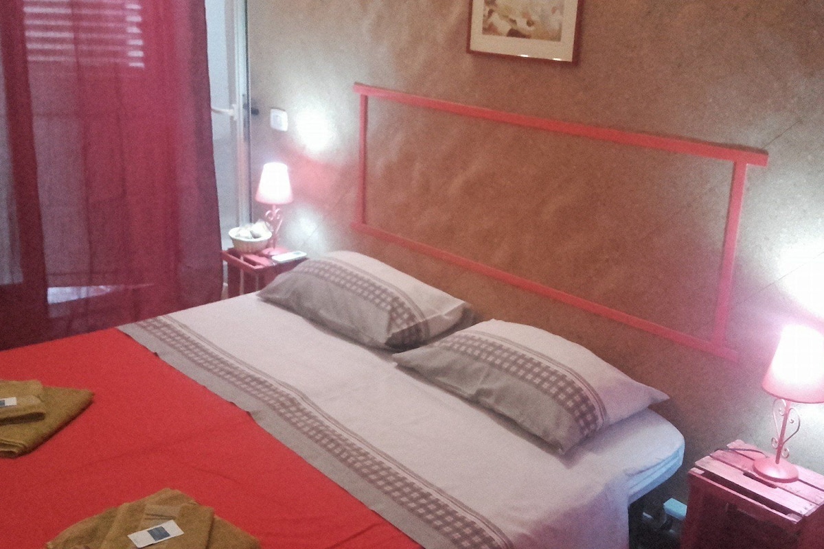 A hotel room with a red bedspread, two pillows, a nightstand with a lamp, and a framed picture on the wall.