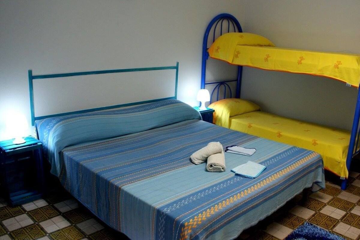 A single bed with a patterned bedspread, a nightstand with a lamp, and a bunk bed with a yellow bedspread.