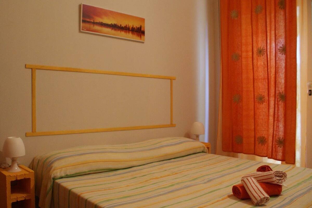 A bedroom with a bed, a nightstand, a lamp, a painting on the wall, and a window with curtains.