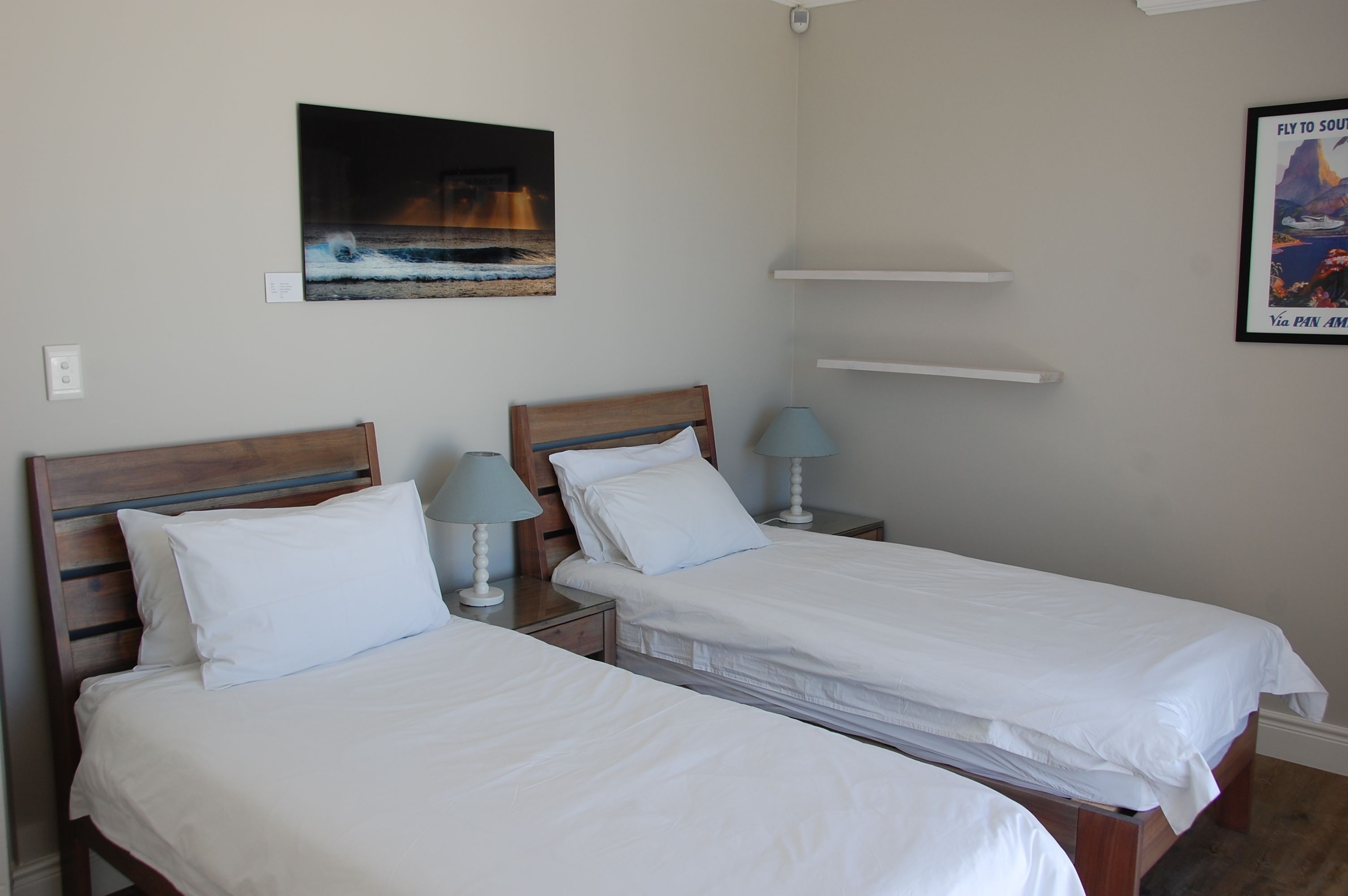 Une chambre d’hôtel avec deux lits simples, des têtes de lit en bois, du linge de lit blanc, des lampes de chevet, une œuvre d’art fixée au mur représentant une scène de plage, et une affiche encadrée accrochée au mur.