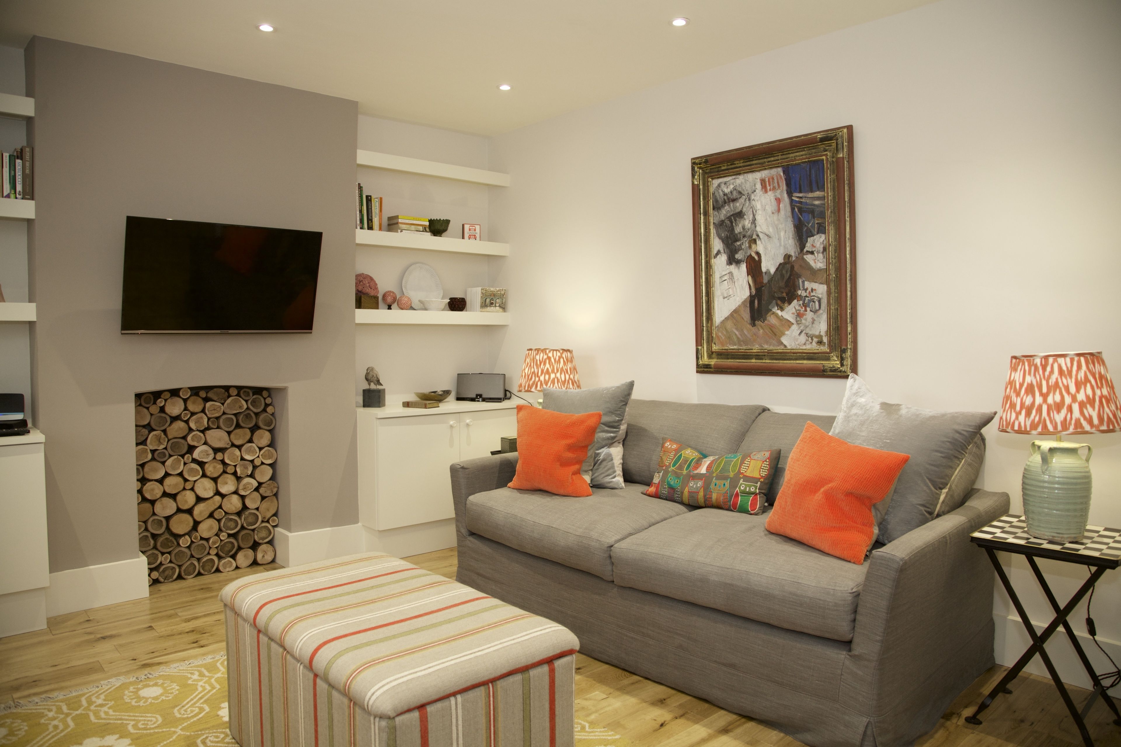 Un salon avec un canapé gris, des coussins orange, un pouf rayé, une cheminée avec des bûches, un téléviseur, des étagères remplies de livres et d’objets décoratifs, et un tableau encadré.