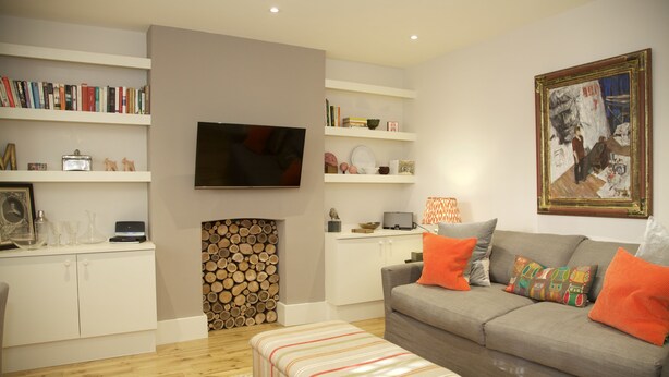 Ein Wohnzimmer mit grauem Sofa, orangefarbenen Kissen, Fernseher, Bücherregalen, Kamin und Teppich mit Muster.