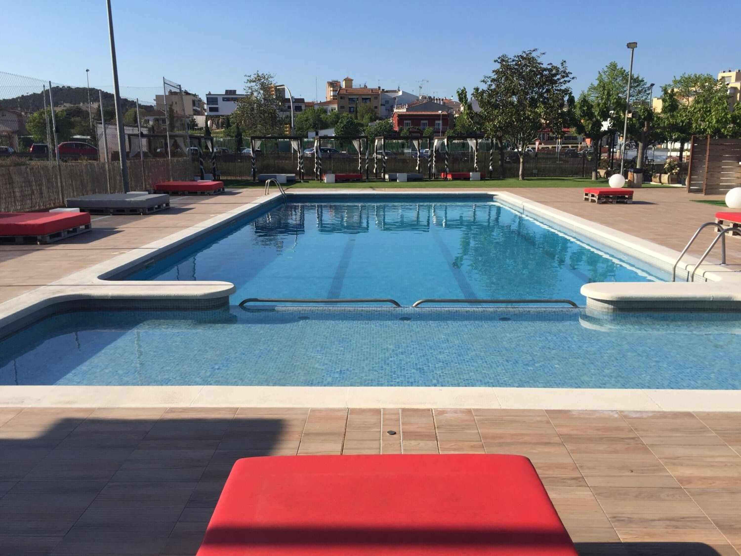 Une piscine rectangulaire à l’eau d’un bleu limpide, entourée d’une terrasse carrelée avec des chaises longues rouges et d’un espace de terrasse en bois.