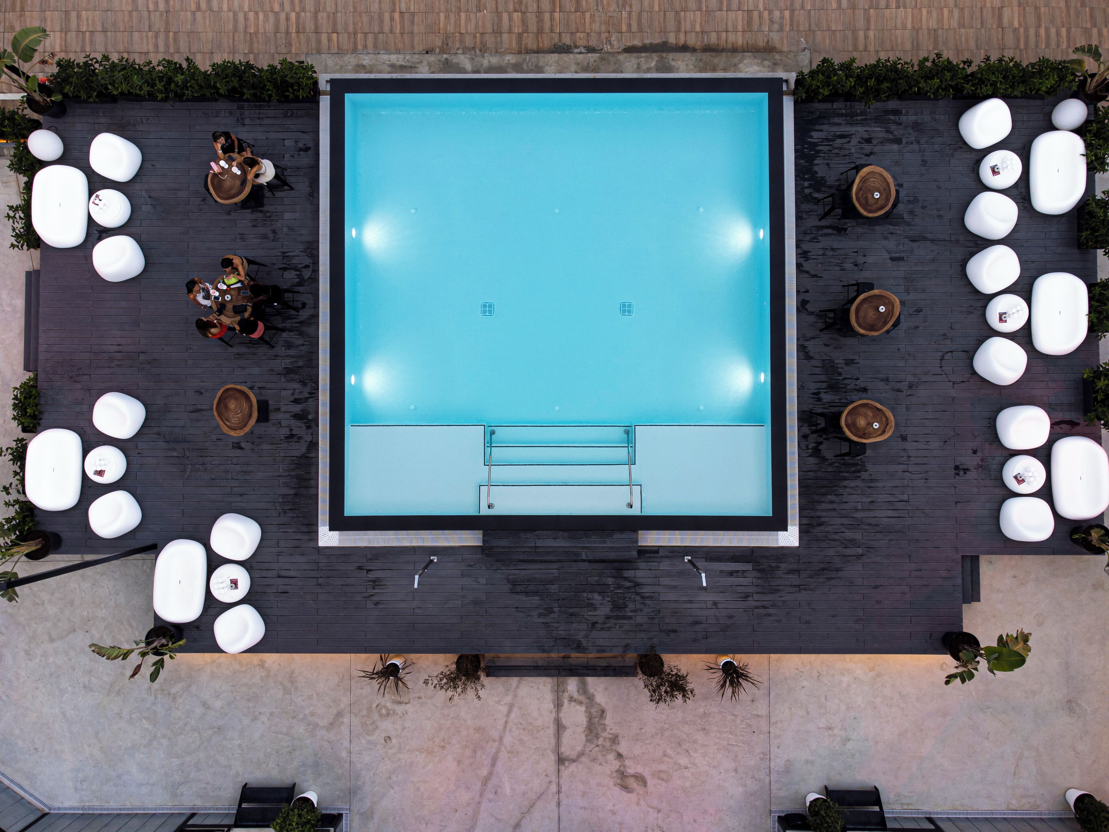 A rooftop pool area with a rectangular pool, surrounded by white lounge chairs and potted plants.