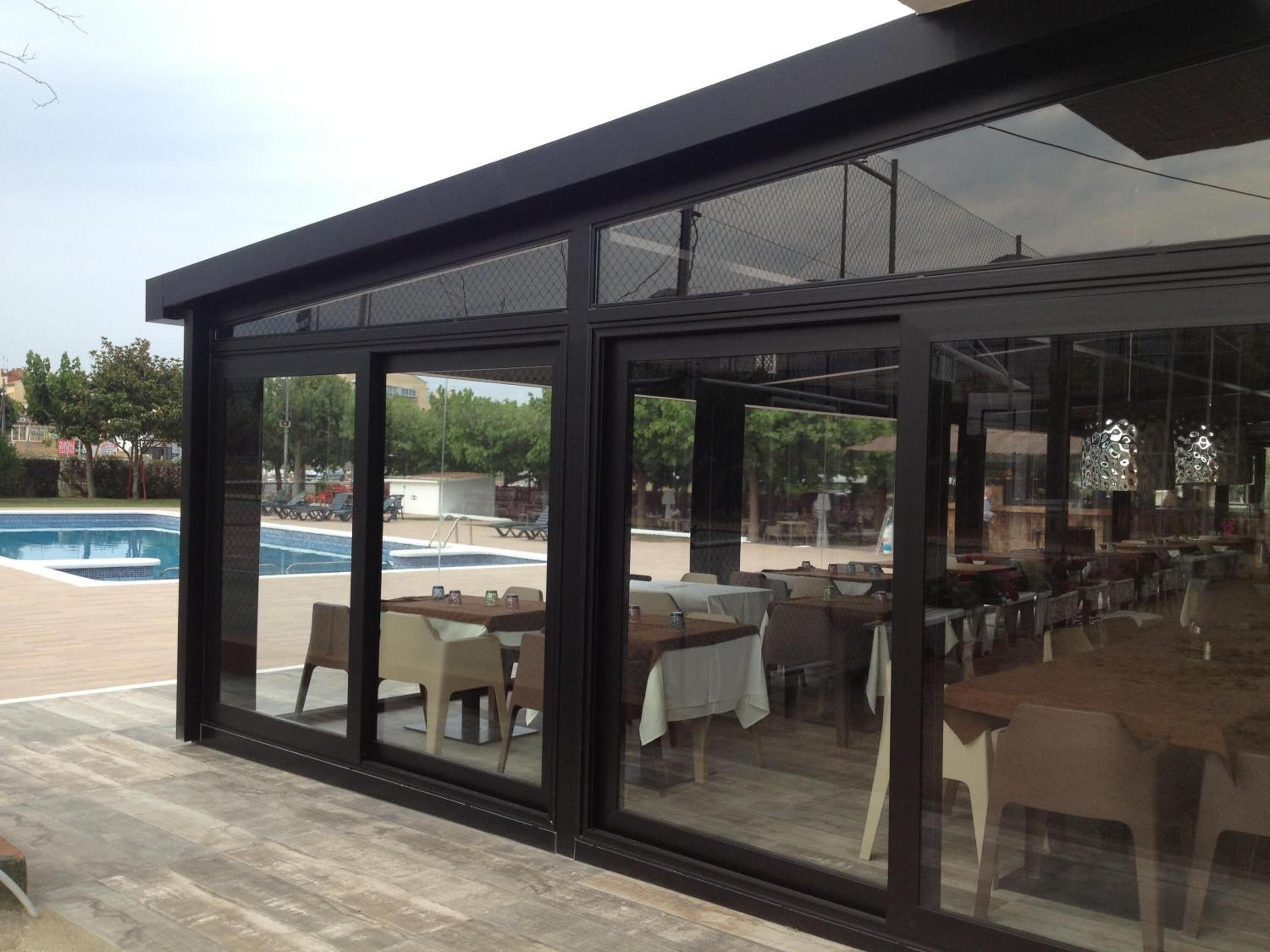 Un área de comedor exterior moderna con paredes de vidrio, una piscina y vistas del paisaje circundante.