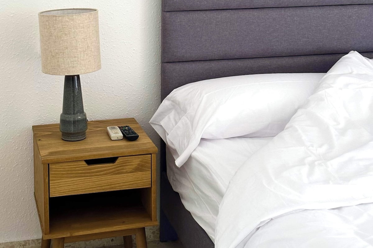 A neatly made bed with white linens, a grey upholstered headboard, a wooden nightstand with a lamp, and a remote control.