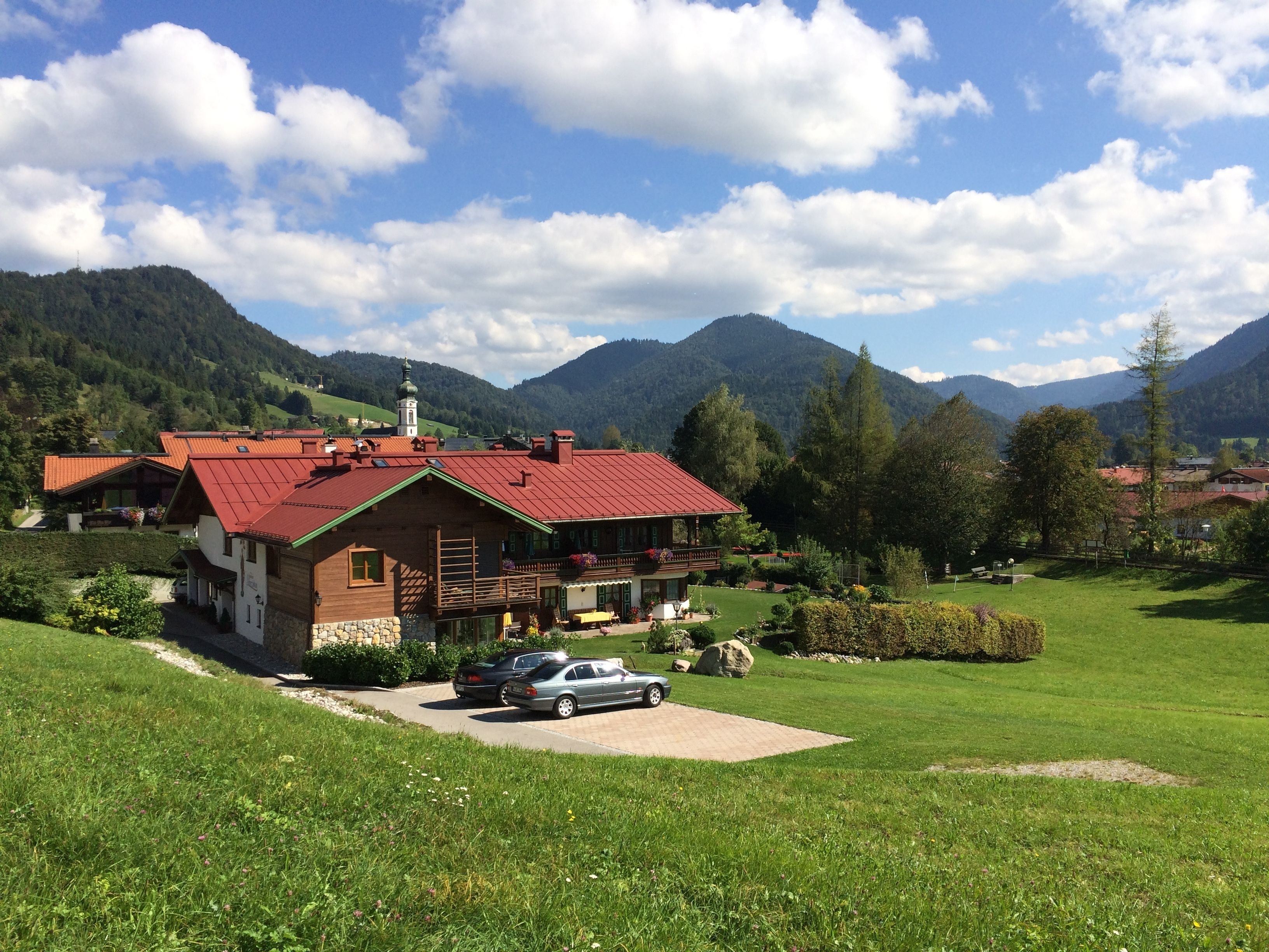 A wooden house with a red roof, surrounded by a well-maintained lawn and a mountain backdrop.