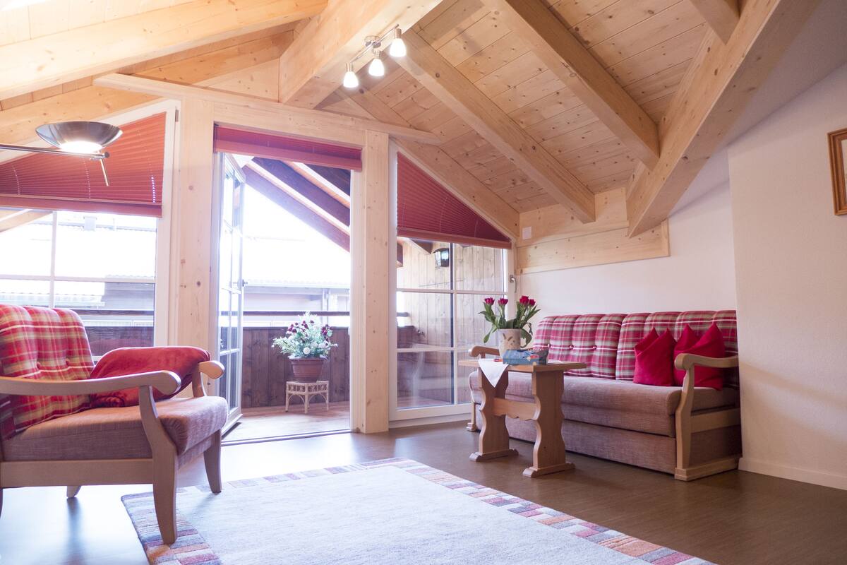 A cozy living room with a wooden ceiling, a plaid sofa, a chair, and a small table with a vase of flowers.