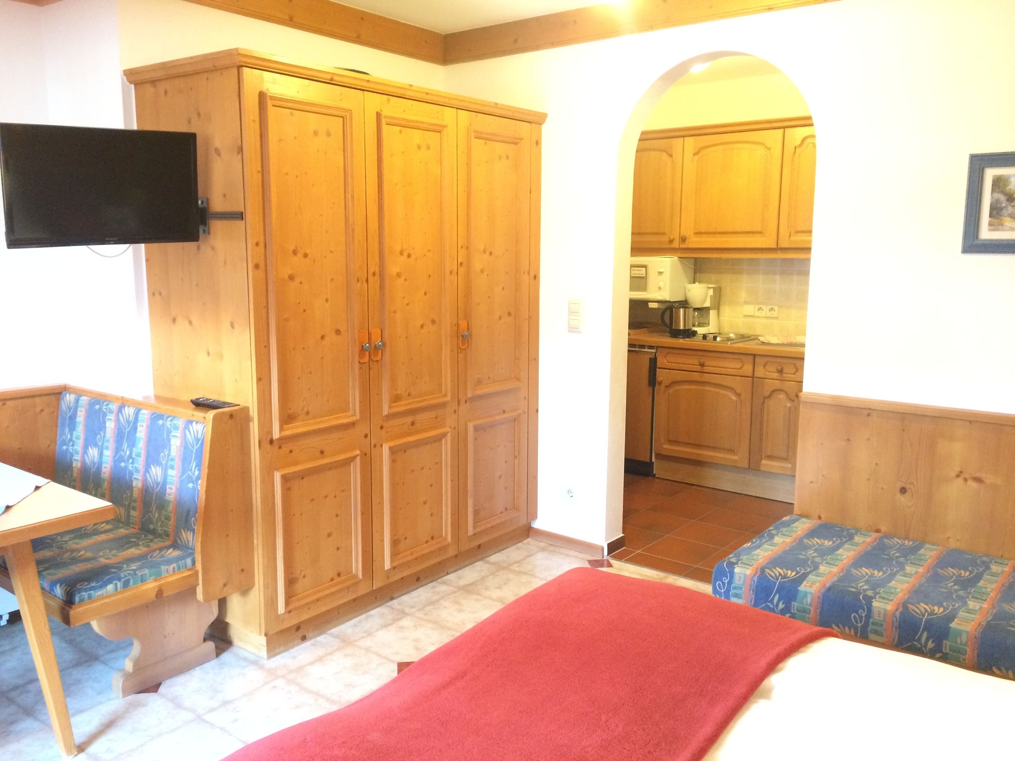 Quarto com armários de madeira, televisão, um banco e uma cozinha.