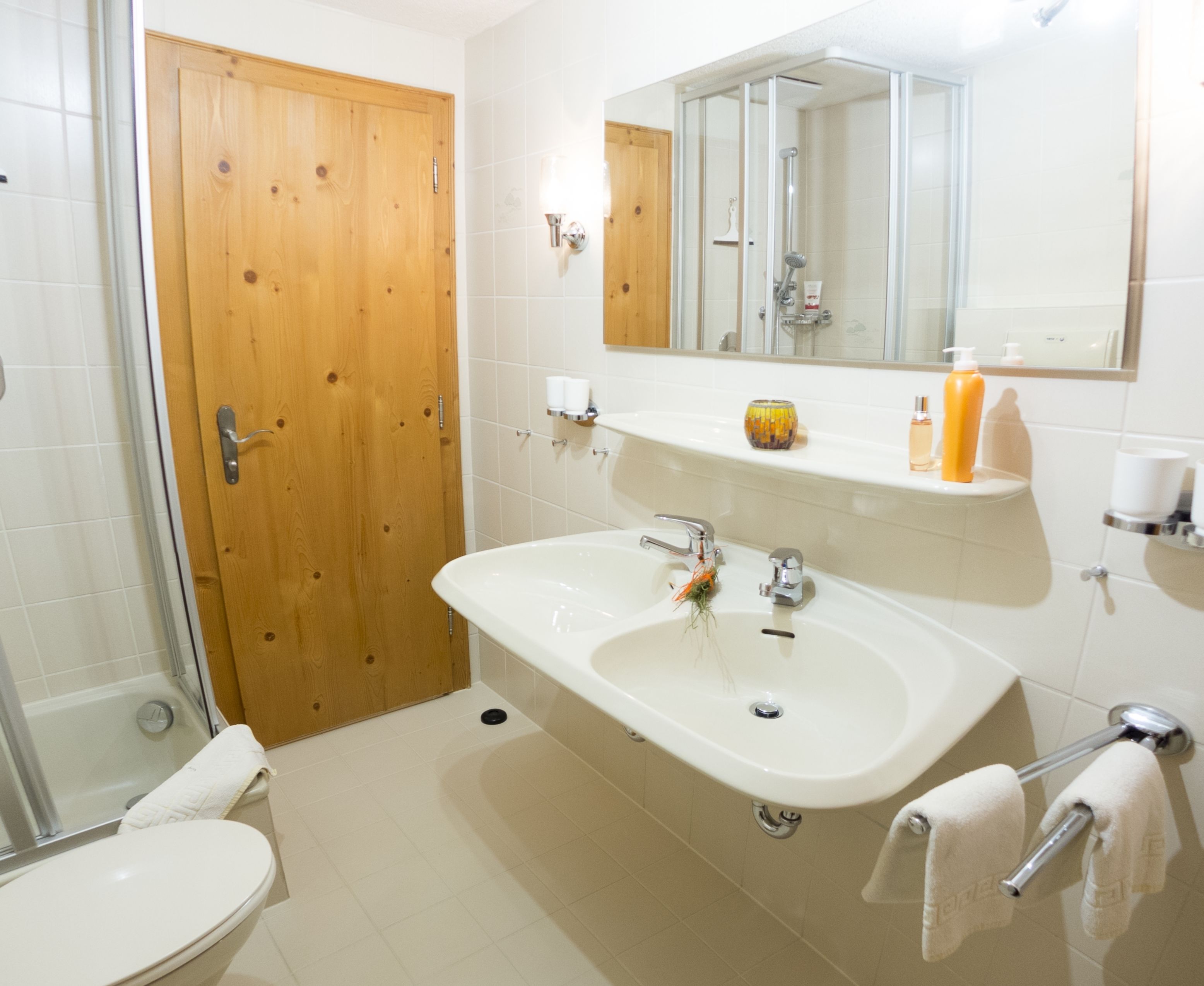 Een badkamer met een houten deur, een witte wastafel, een spiegel en een douche.