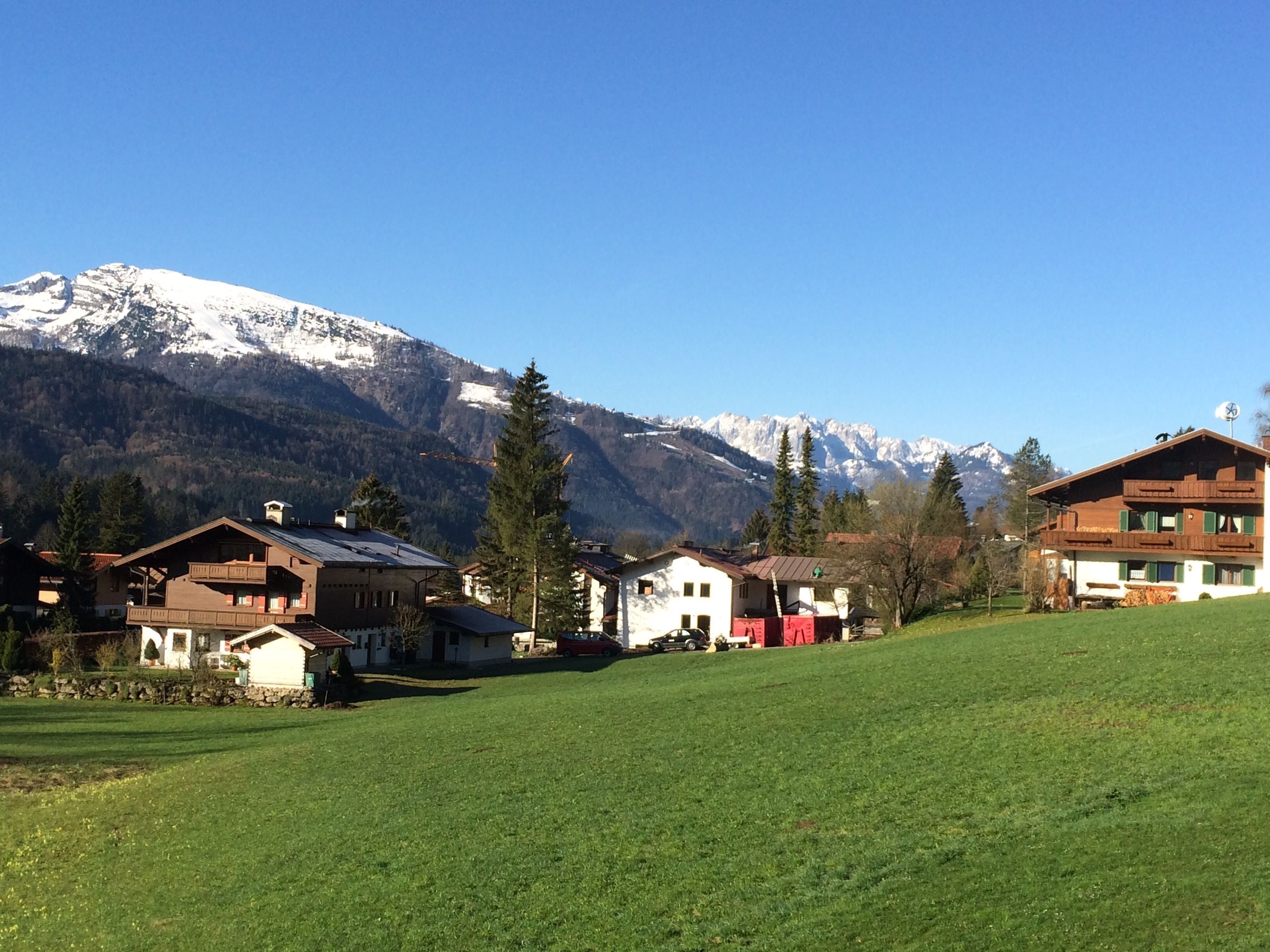 Ein Bergdorf mit traditionellen Holzhäusern, einer grünen Wiese und schneebedeckten Bergen im Hintergrund.