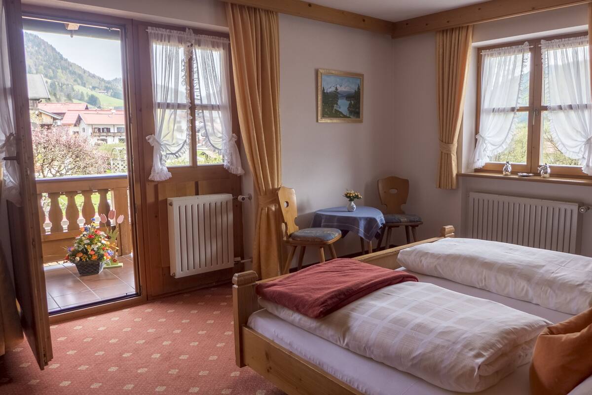 A room with two beds, a table with chairs, a window with curtains, and a view of a mountain landscape.