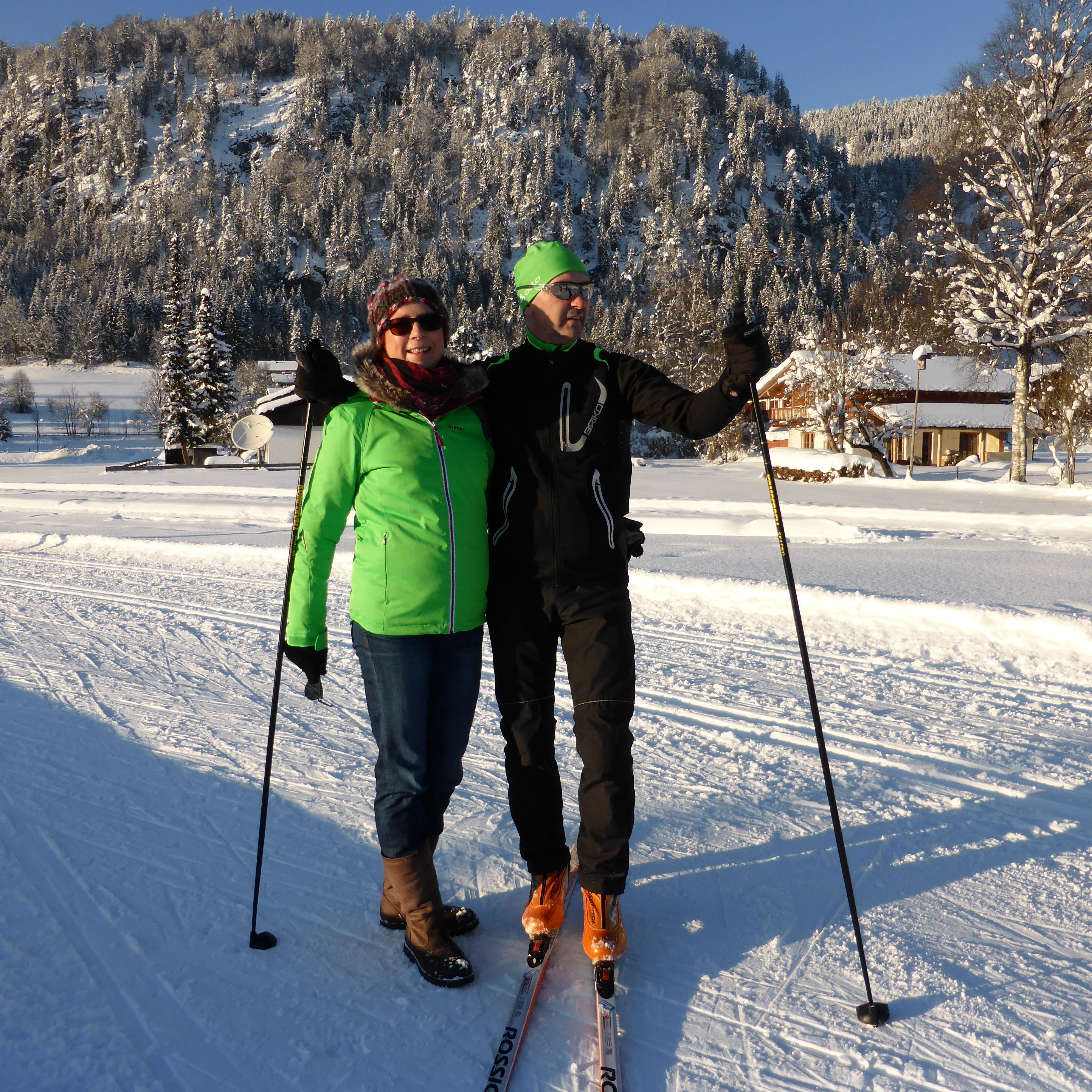 Two people on skis in a snowy landscape with a cabin in the background.