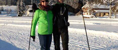 Two people on skis in a snowy landscape with a cabin in the background.